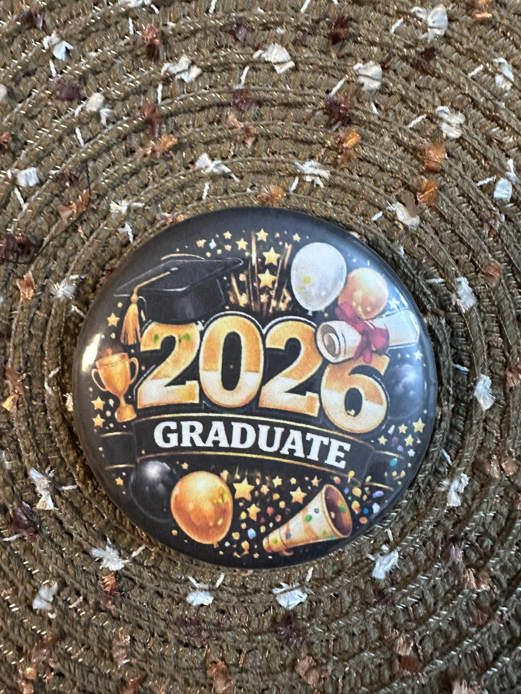 Celebrate the Class of 2026 with this bold and festive 2.25" graduation button pin. Featuring a striking gold “2026 Graduate” design with balloons, stars, and celebratory accents, this pin is perfect for graduates and supporters alike. Lightweight with a durable plastic back, it’s comfortable to wear and makes a great keepsake for graduation ceremonies, parties, and photos.