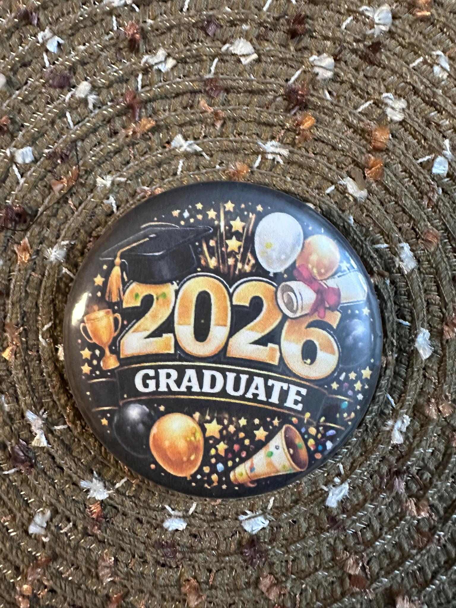 Celebrate the Class of 2026 with this bold and festive 2.25" graduation button pin. Featuring a striking gold “2026 Graduate” design with balloons, stars, and celebratory accents, this pin is perfect for graduates and supporters alike. Lightweight with a durable plastic back, it’s comfortable to wear and makes a great keepsake for graduation ceremonies, parties, and photos.